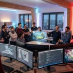 Diverse French game developers collaborate in a modern studio, viewing a holographic game world, code, and 3D models on screens.
