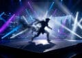Dynamic armored silhouette emerging from a digital void on a futuristic stage, lit by vibrant blue, purple, and white spotlights.