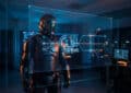 Armored figure in a futuristic command center views a holographic 'PAUSED' display with data connections, bathed in blue light.