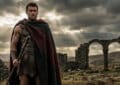A muscular man in ancient attire, red and grey cloak, stands confidently in rugged ruins under a dramatic, light-shafted sky.
