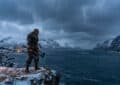 Photorealistic image of a Viking warrior in armor on a snowy cliff, holding axes, overlooking an icy fjord and mountains at dusk.
