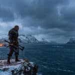 Photorealistic image of a Viking warrior in armor on a snowy cliff, holding axes, overlooking an icy fjord and mountains at dusk.