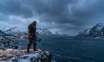 Photorealistic image of a Viking warrior in armor on a snowy cliff, holding axes, overlooking an icy fjord and mountains at dusk.